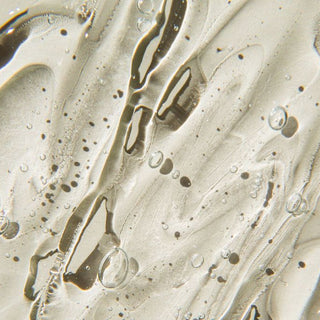 Close-up of a translucent, glossy cosmetic cream or gel swirled on a light beige background, showing a clear, slightly viscous texture with small dark specks and reflective highlights.